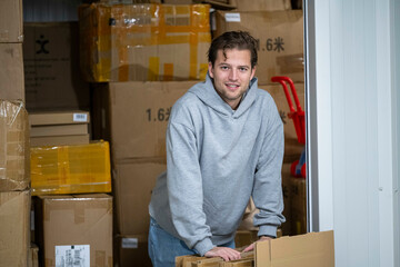 A young man in a gray hoodie leans on a stack of cardboard boxes in a storage area, surrounded by several large parcels. He is smiling and looks relaxed, Molenhoek, Netherlands