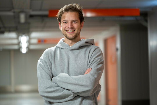 A smiling young man in a gray hoodie stands confidently in an indoor setting with soft lighting and an industrial background, Molenhoek, Netherlands
