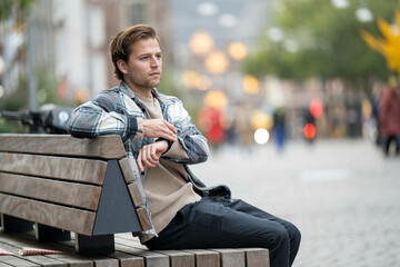 Young man with a smartwatch sits on a wooden bench in an urban setting, wearing a plaid shirt and jeans. He appears thoughtful, with blurred city lights, Nijmegen, Netherlands