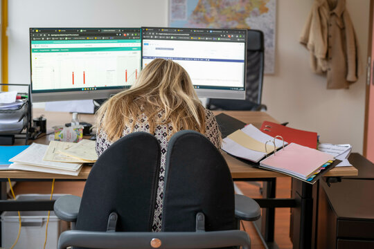 A woman with long blonde hair sits hunched over at a desk cluttered with papers and files, facing two computer monitors displaying data charts and spreadsheets, Molenhoek, Netherlands