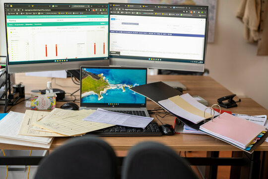 A cluttered office workspace with dual computer monitors displaying graphs and spreadsheets, a laptop featuring a landscape, and scattered documents, folders, and stationery, Molenhoek, Netherlands