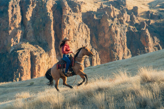 A woman in a red shirt and cowboy hat rides a horse through a golden grass hillside with rugged cliffs in the background, Idaho, USA
