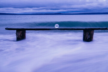 A serene seascape at dusk with a long, weathered wooden bench partially submerged in swirling ocean waves, Norwegian Point, WA, USA
