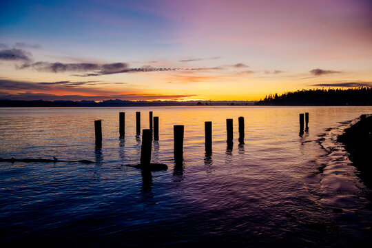 A serene sunset over calm waters with silhouetted wooden posts emerging from the lake. The sky transitions from deep orange near the horizon to a rich purple higher up, Norwegian Point, WA, USA