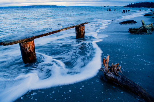 Ocean waves gently wash over a rustic wooden bench embedded in the sandy shoreline, Norwegian Point, WA, USA