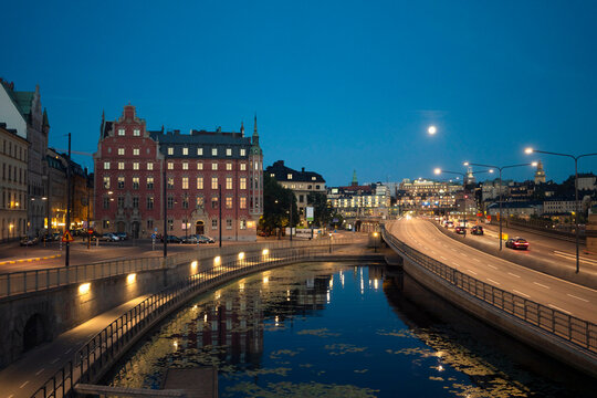Twilight cityscape with historical buildings reflected in the calm waters of a canal in Gamla Stan, under a clear sky with a bright full moon, Stockholm, Sweden