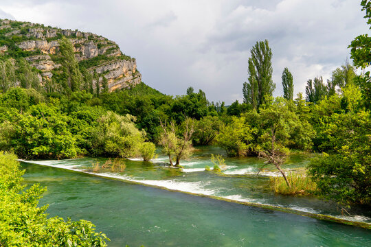 Tranquil river flowing through a lush landscape with overcast skies and towering cliffs in the background, Krka waterfalls at the national park, Croatia