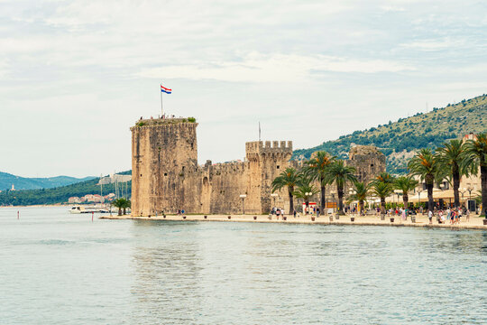 Historic Kamerlengo castle towers over a tranquil Adriatic seafront promenade lined with tourists and lush greenery under a clear sky, Trogir, Croatia