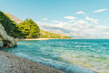 Serene Artina beach with clear turquoise water of Adriatic sea, lush green trees, and a mountain backdrop under a blue sky with clouds, Croatia