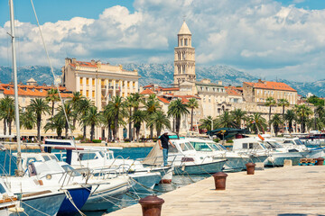 Marina with boats moored along the waterfront, with a historic bell tower of St domnius Cathedral and buildings against a backdrop of mountains and a cloudy sky, Split, Croatia