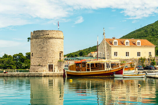 A traditional wooden boat moored beside a historic stone tower along a calm waterfront with clear skies and a hill in the background, Mali Ston, Croatia