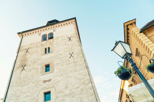 Fototapeta White medieval Lotr??ak Tower with a green roof under a clear blue sky, flanked by historic buildings and street lamps adorned with hanging plants, Zagreb, Croatia