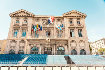 Classical architecture building of city hall adorned with French flags under a clear blue sky, with rows of blue seating in front, Marseille, France