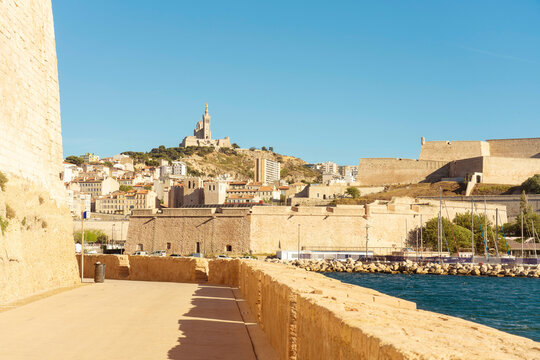 Sunny day view of a historic Fort Saint-Jean and Citadelle de Marseille by the sea with a cityscape and hilltop Basilica of Notre-Dame-of-la-Garde in the background, Marseille, France