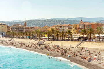 Sunny beach scene with a crowd of people enjoying the shore, the turquoise sea, and a backdrop of a colorful coastal town of Nice, France