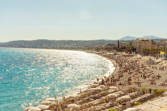 Sunny beach scene with a crowd of people enjoying the shore, the turquoise sea, and a backdrop of a colorful coastal town of Nice, France