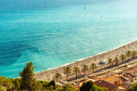 High-angle view of the bustling beach of Nice city in the french riviera with sunbathers, a parasail in the distance, and crystal-clear turquoise waters.  