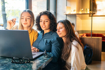 Three women focused on a laptop screen while sitting at a table with warm lighting in the background, Berlin, Germany