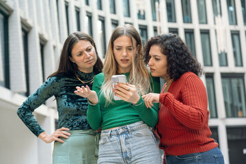 Three women looking at a smartphone with confused expressions in an urban setting, Berlin, Germany
