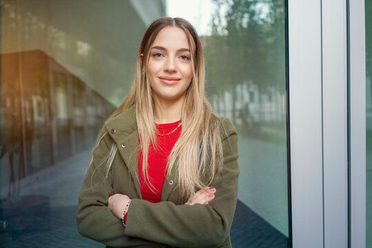Smiling young woman in a green jacket and red top standing with arms crossed near a glass building, Berlin, Germany