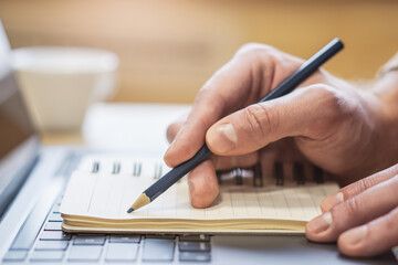 Sharp view of a man's hand transcribing in a notepad on a cutting-edge laptop, with a defocused backdrop