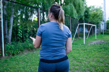 A woman in athletic wear plays badminton alone in a park. The shuttlecock is airborne, and a soccer goal is visible in the background.