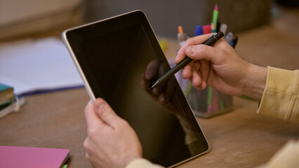 Close-up of hands holding tablet and stylus pointing at blank screen with organized desk setup,...