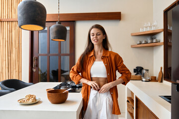 Young woman in a stylish kitchen preparing healthy food while wearing an orange shirt and white pants conveying a sense of wellness and lifestyle