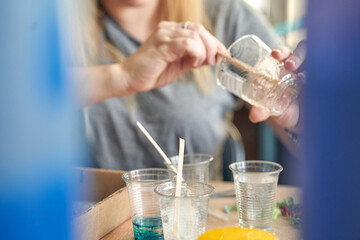 Female adult pouring liquid into clear plastic cups with stirring sticks in creative workshop setting.