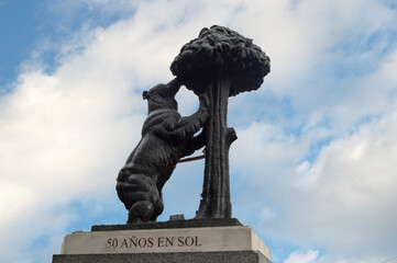 Statue of the bear and the strawberry tree of Madrid against a cloudy sky. On the pedestal is written in Spanish "50 years in the sun"