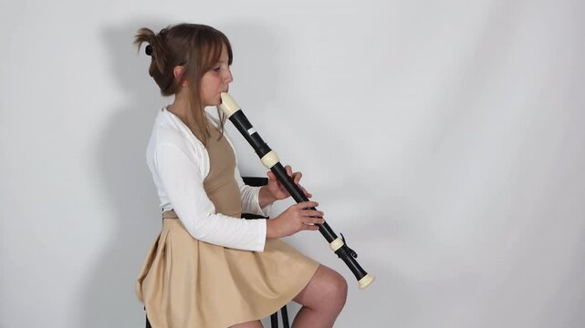 Girl playing, practicing the flute, playing a melody on a musical instrument, wooden pipe, sitting on black chair on white background. Out-of-school children activities, lessons