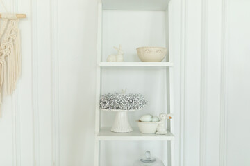 Wooden shelf on the wall with easter decorations, festive wreath, ceramic plates and utensils. Cozy interior design composition of easter living room interior with bunny sculpture and eastrer eggs. 