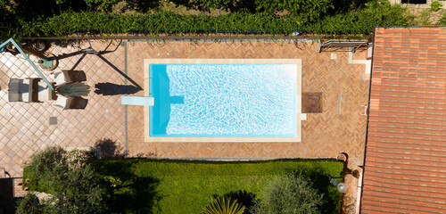 Aerial perpendicular view of a rectangular pool outside a modern house with a garden. The pool has a diving board but is empty.