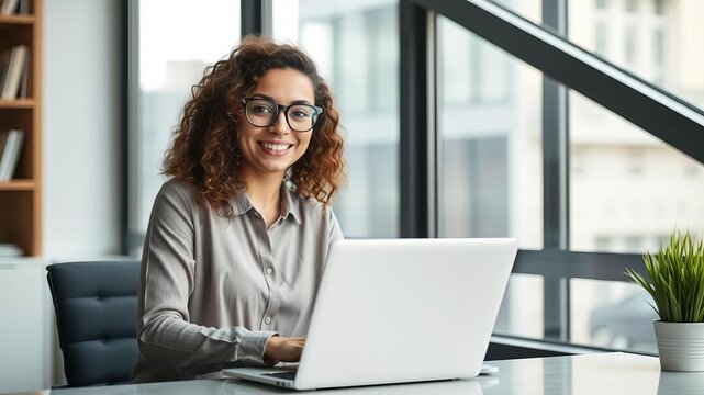Portrait of a young beautiful woman with curly hair and glasses standing in an office, smiling as she looks out the window while working on her laptop, curly hair, freelancer, female