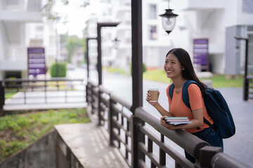 Young woman with a backpack and books enjoying a coffee break on a university campus, smiling brightly while gazing into the distance, surrounded by greenery and modern architecture