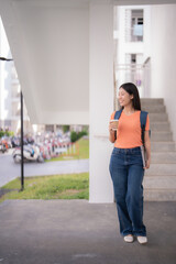 Young Asian student walking outside her university building, sipping takeaway coffee and carrying a laptop, enjoying a relaxing break in the vibrant campus atmosphere