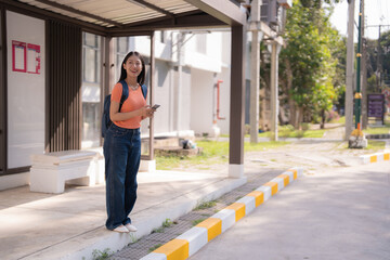 Happy asian university student is standing at bus stop, holding smartphone and waiting for bus,...