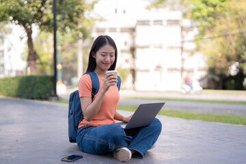 Young woman is sitting with crossed legs on the ground in a park, drinking coffee and working on a laptop, with a backpack and a smartphone next to her