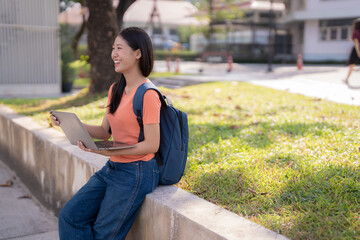 Happy young asian woman with backpack using laptop sitting on a low wall in university campus,...