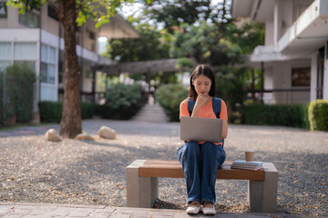 Fototapeta premium Concentrated young Asian female university student wearing backpack using laptop and thinking about project while sitting on bench at campus during break
