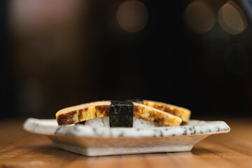 Tamago sushi wrapped in seaweed, beautifully served on a ceramic plate with a blurred, dramatic background.