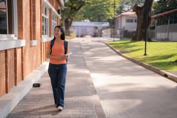 Happy Asian student walking along a university campus, holding books and smiling brightly, embracing her educational journey under the warm sun on a beautiful day