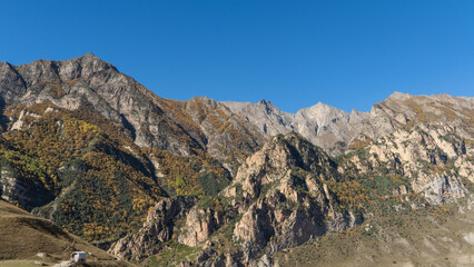 Fototapeta premium Chegem Gorge. Kabardino-Balkaria. El-Tyubu. Unique rocky ridge with steep cliffs against blue autumn sky.Rocky mountains with trees growing on their slopes glow in morning sun.