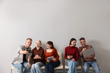 Fototapeta premium Group of people using different gadgets on chairs near light grey wall indoors. Modern technology
