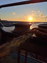 Sunset view from a harbor with a rusty bollard and mooring rope in the foreground, highlighting maritime and industrial elements