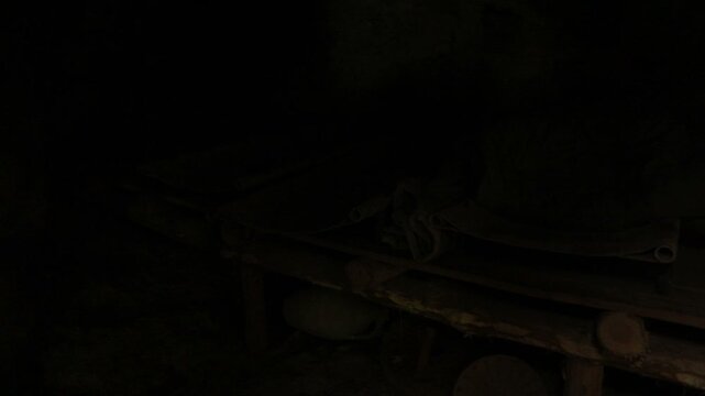 A dim, cave-like space shows rudimentary cots made of wooden logs. Two cots are canvas and a sleeping bag sits on one. An old jar rests beneath. Scene suggests a rustic shelter.