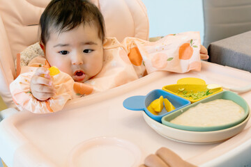 A baby with infantile strabismus. Strabismus loss of eye parallelism in children. Baby eating a nutritious and healthy breakfast in a highchair tries to focus with his eyes on the fruit in his hand.