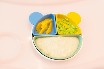 top view of fun children's plate with baby food on pink table with copy space. Food consists of mashed potatoes, avocado and orange fruit wedges. concept: healthy and colorful baby food.