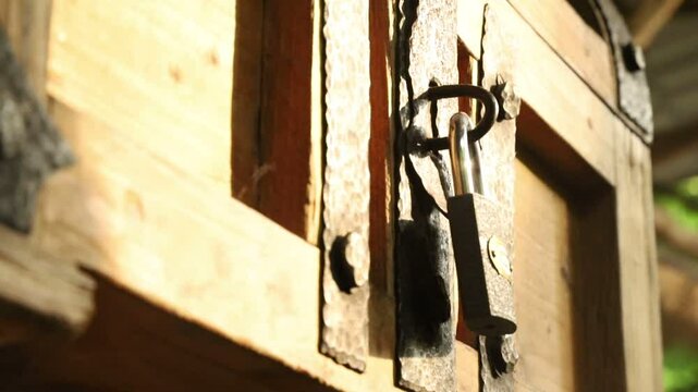 Close-up of a wooden chest or door with rustic metal hinges and a hasp secured by a padlock. The wood is aged with visible grains, illuminated by sunlight creating contrasting shadows. 