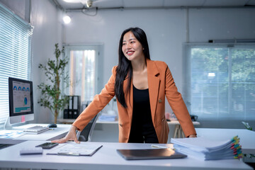 A woman in an orange jacket stands in front of a desk with a computer monitor